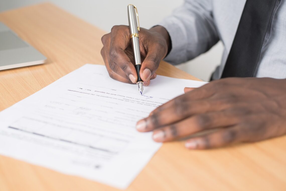 close-up of a document with two hands, one of which is signing the bottom of the page.