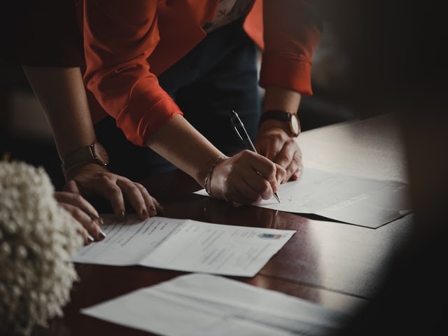 Close up of two people's arms signing documents spread out on a table.