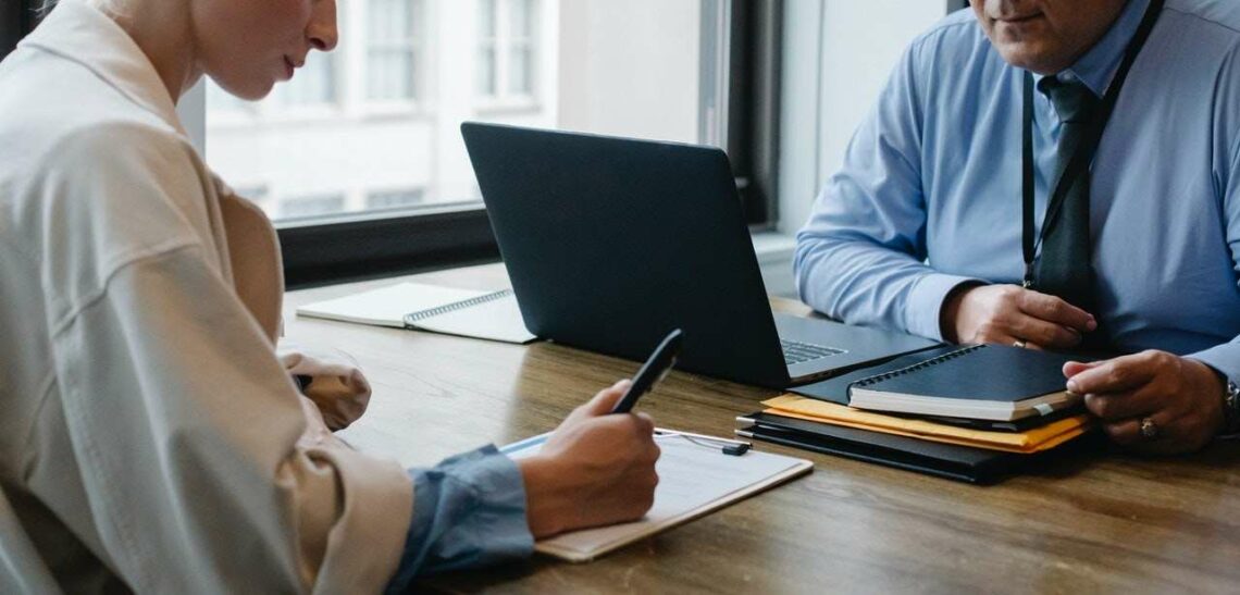 Two people doing paperwork sitting across a desk from one another.