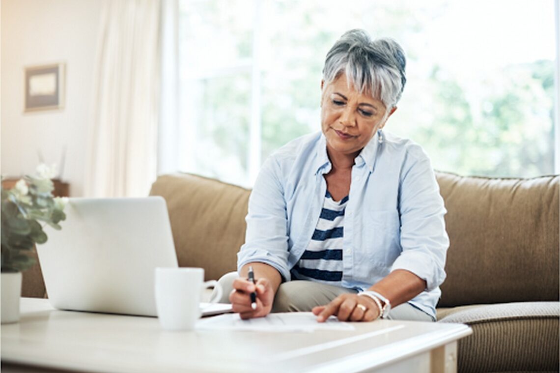 Senior woman sitting in front of a laptop writing on paper.