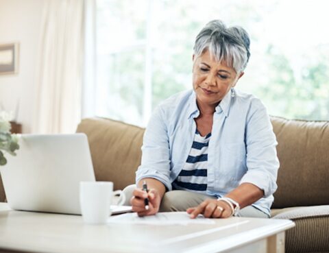 Senior woman sitting in front of a laptop writing on paper.